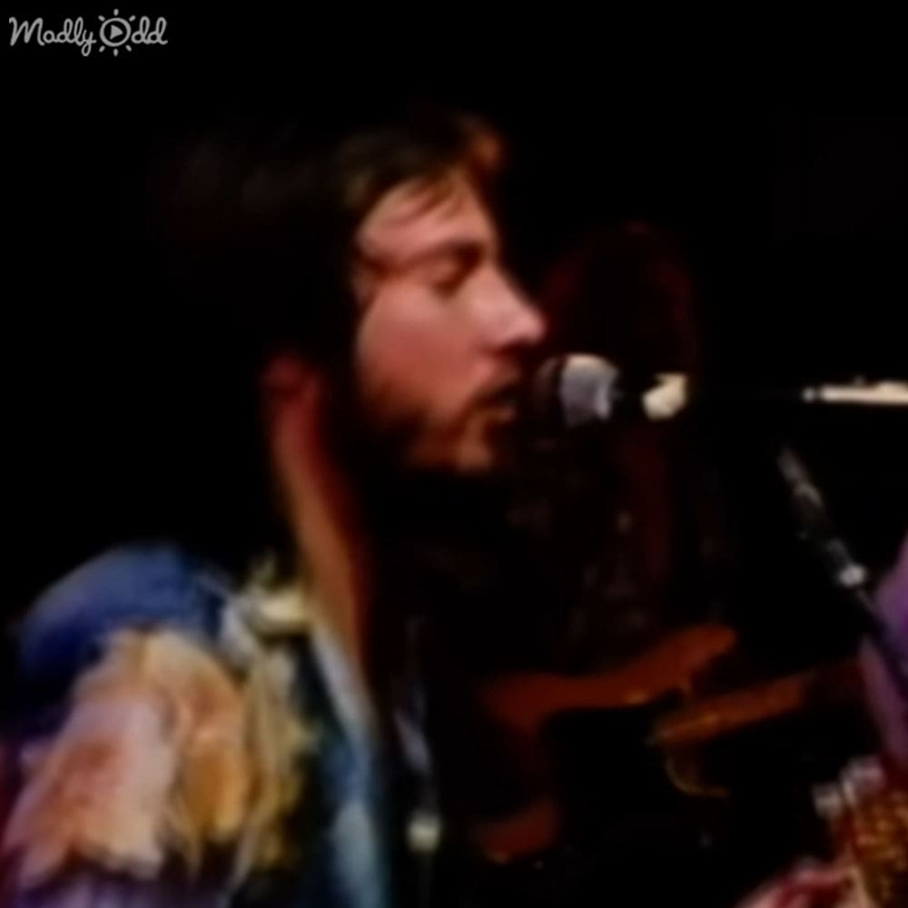 A young adult male singer performs on stage at a music venue, wearing a light-colored, loose-fitting floral shirt with a collar and long sleeves.