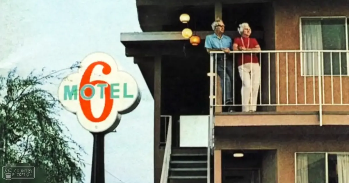 An older adult male singer wearing a light blue collared shirt and white pants, standing outside a Motel 6 with green trees in the background during daytime in spring or summer.
