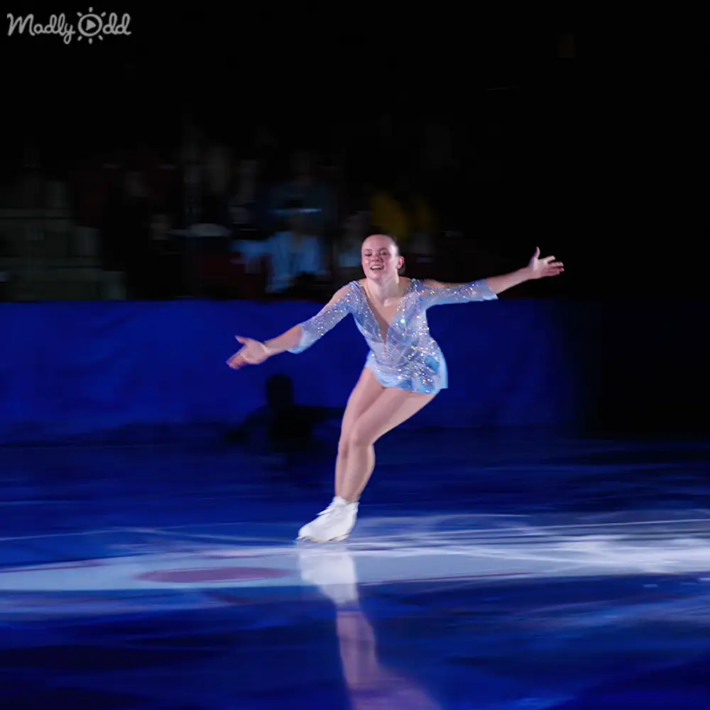A young adult female figure skater in a sparkly light blue dress performing at an indoor ice rink with artificial lighting and smooth ice.