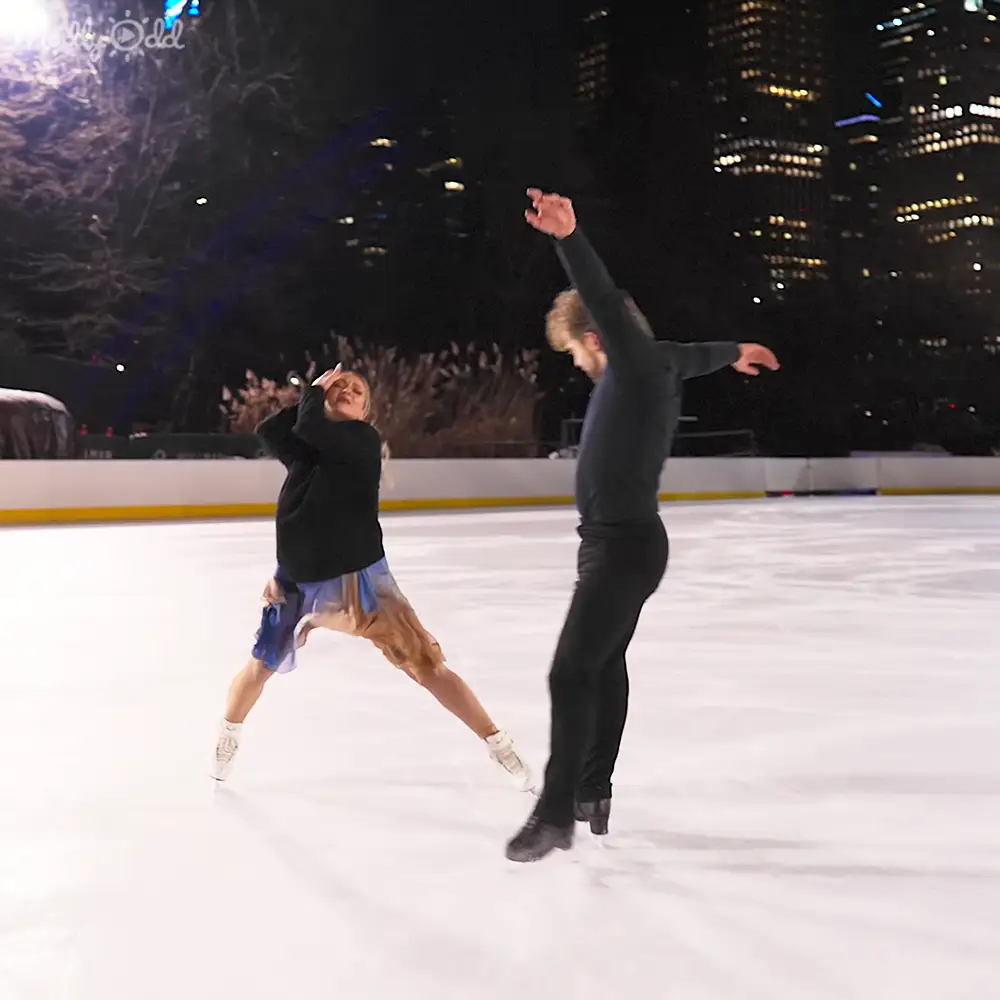Two people ice skating at an outdoor rink surrounded by a cityscape at night, with buildings lit up in the background.