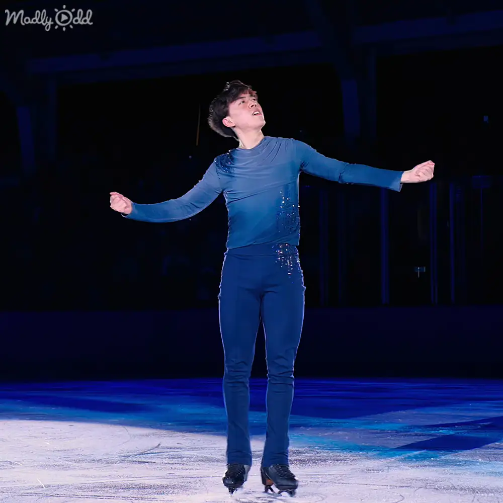 A young adult male figure skater in a form-fitting blue performance outfit with sparkly embellishments, skating on an indoor ice rink under spotlights at night.