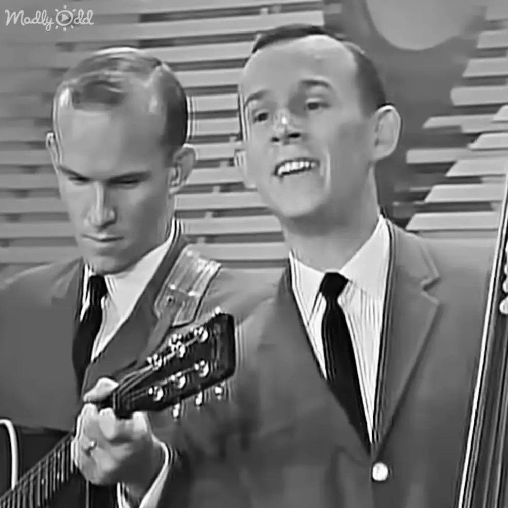 A musical performance scene with young adult male performers wearing dark tailored suits against a patterned backdrop, likely in an indoor television studio or concert venue.