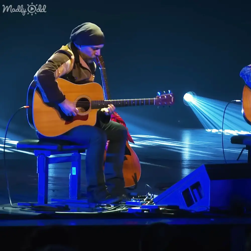 A male adult musician in a beanie hat, patterned jacket, long-sleeved shirt, jeans, and boots performs live on stage with musical instruments and professional stage lighting.