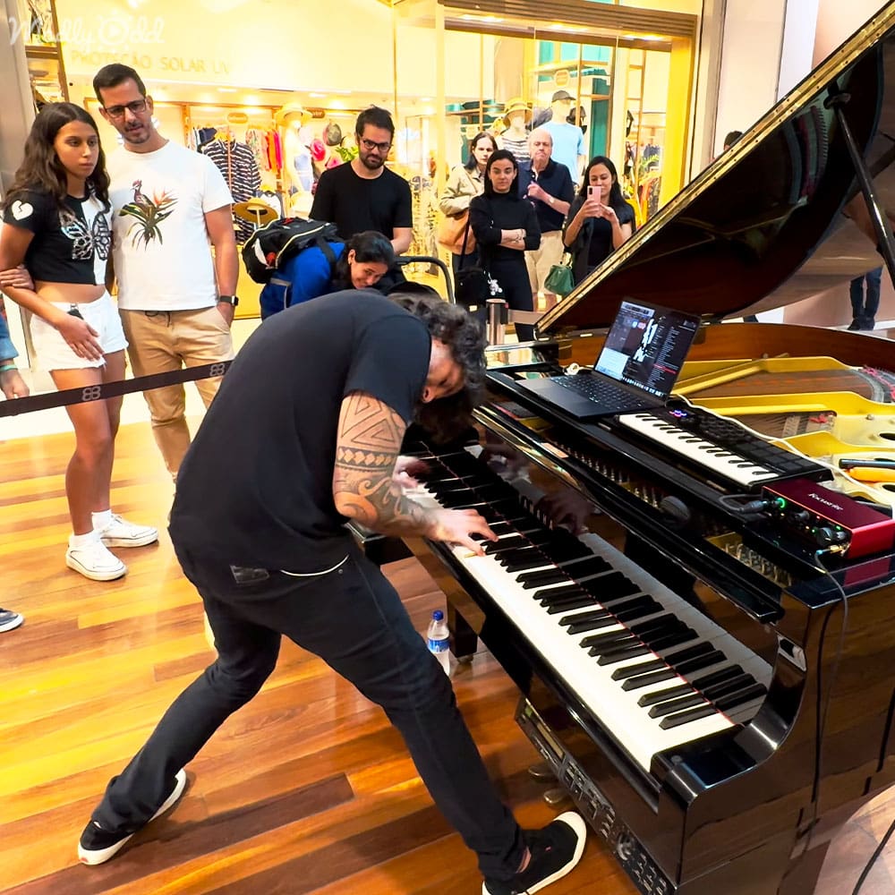 Adult male playing piano surrounded by shoppers in a well-lit shopping mall with various clothing racks and displays visible in the background.
