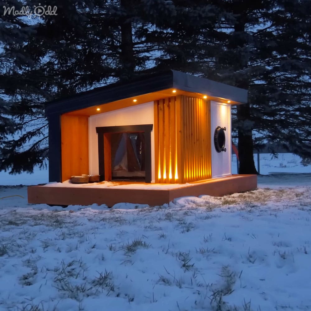 A small wooden cabin with a fireplace, circular window, illuminated interior and exterior, set against snow-covered ground in winter at dusk or early evening.