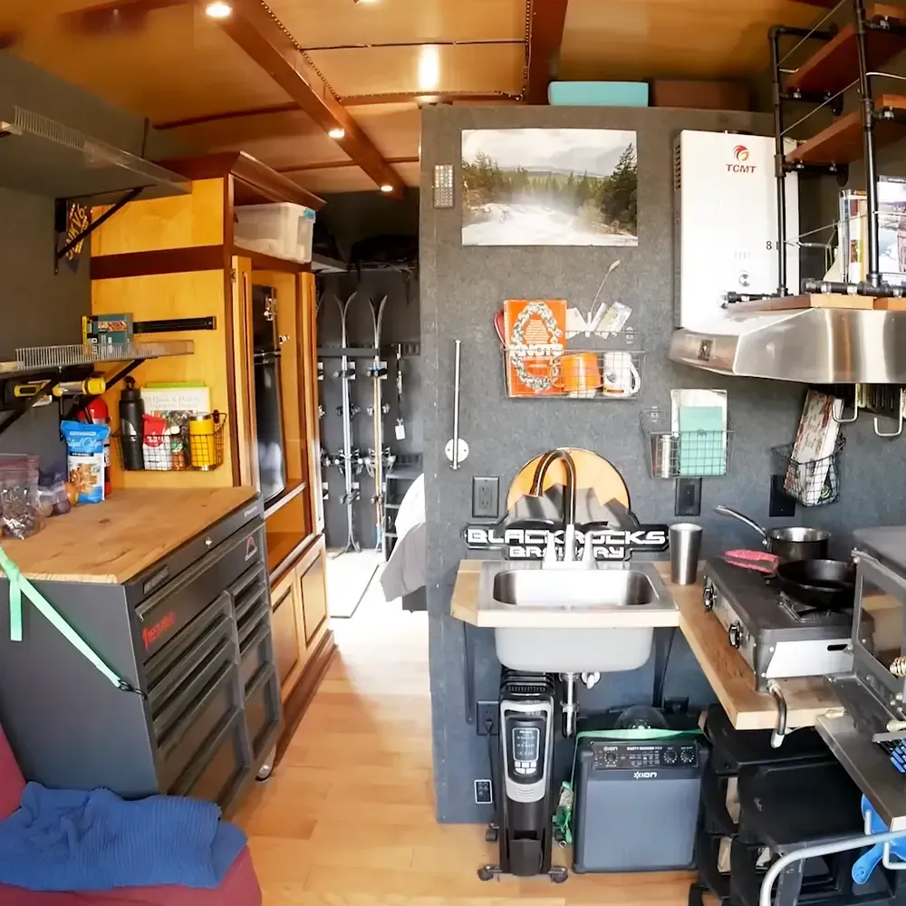 Compact kitchen setup in an RV or camper van with a sink labeled 'BLACKROCKS BREWERY', small stove, coffee maker, utensils, mugs, and ski poles. Sunny day indoors during spring or summer.