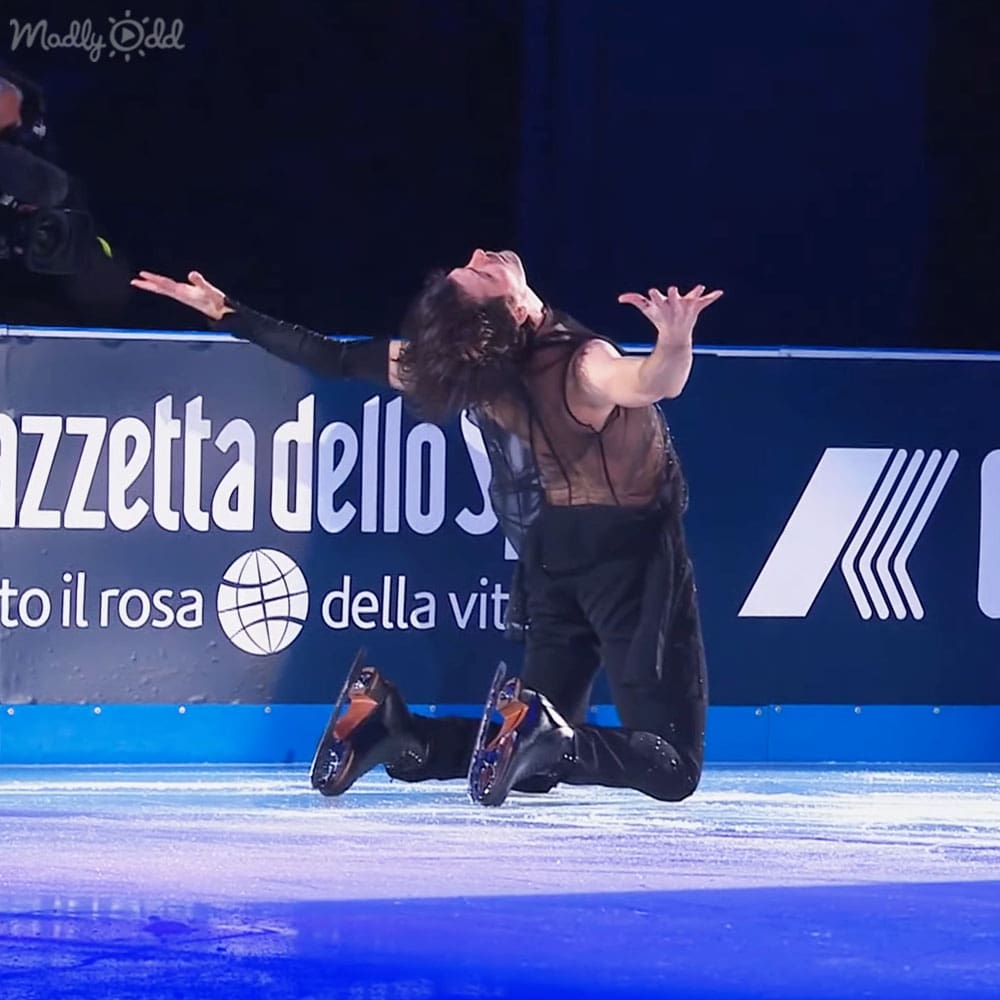 A young adult female figure skater in a black sleeveless top with sheer panel and orange-accented ice skates performing on an indoor ice rink under focused lighting, with a banner displaying 'azzetta dello sport' behind her.