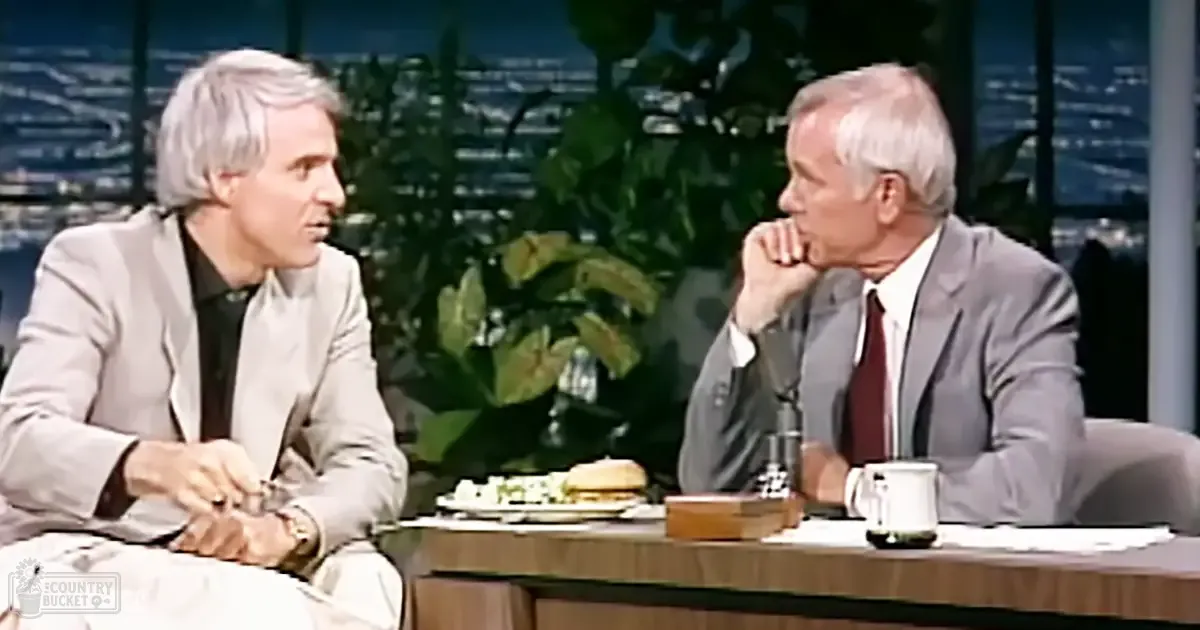 A talk show set with two middle-aged men, one wearing a light gray suit and the other in a gray suit with a red tie, both seated at a wooden table with food and objects. The backdrop features city lights and greenery.