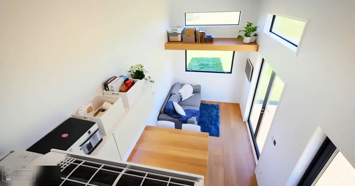 A modern living space with a gray sofa, blue rug, wooden coffee table, floating shelf holding books and a potted plant, kitchenette area with white storage bins, two windows letting in natural light.