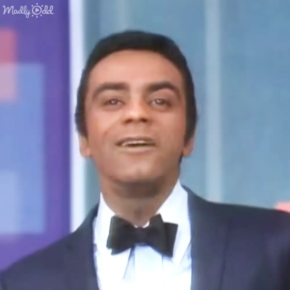 A middle-aged adult male wearing a black tuxedo with bow tie, standing against blue curtains in an indoor studio setting.