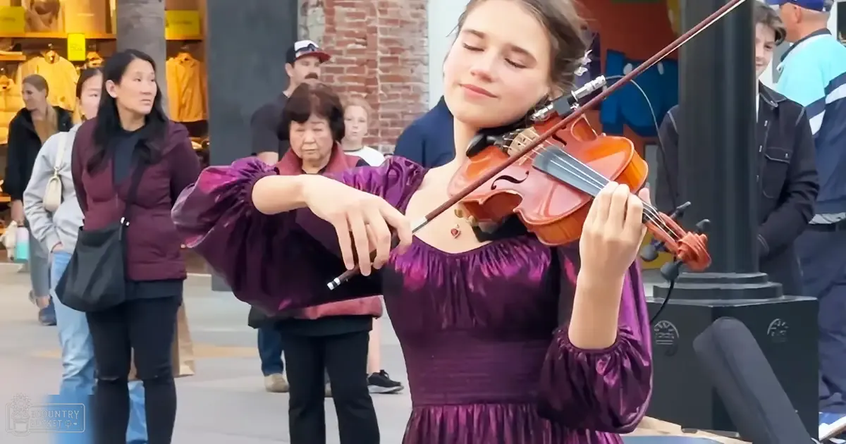 A young adult female playing a violin outdoors, wearing a purple dress with puffed sleeves. Microphone attached to her dress suggests a performance setup.