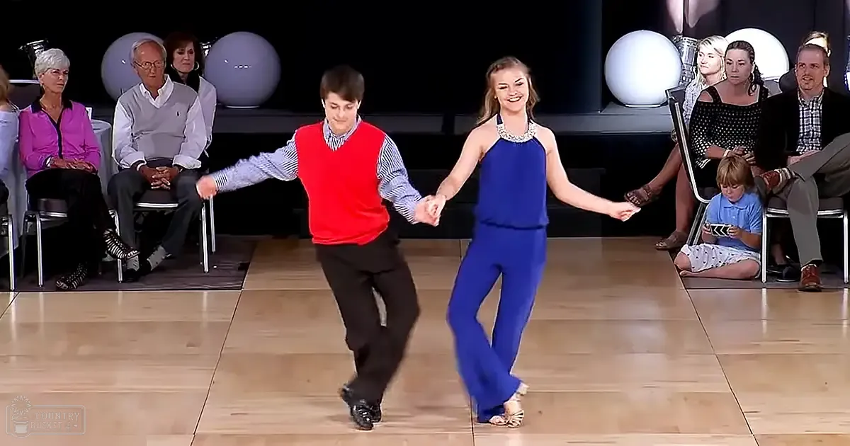 A dance couple performing on stage in a studio setting with seated adults and decorative spherical objects in the background.