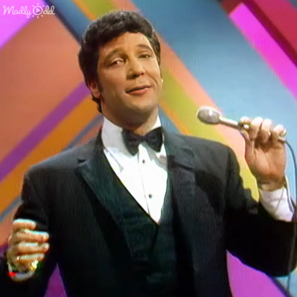 Adult male singer wearing a well-fitted black tuxedo with white shirt and black bow tie, holding a microphone and drink in an entertainment setting with vibrant geometric backdrop.
