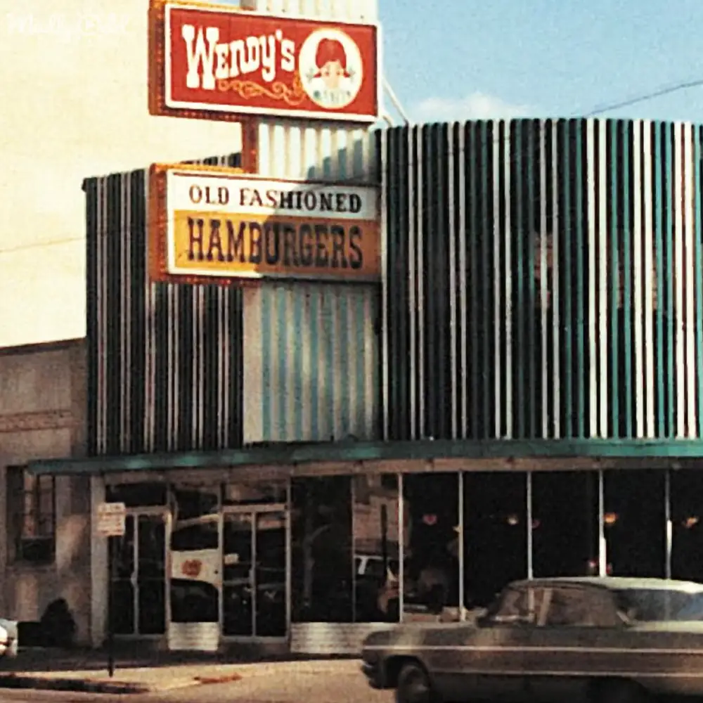 Wendy's restaurant with 'Old Fashioned Hamburgers' sign in urban mid-20th-century setting; vintage car parked, clear blue sky above