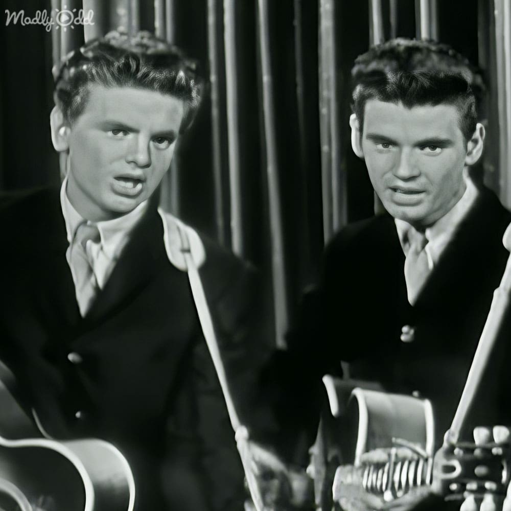 Two young adult male performers in formal suits holding guitars against a backdrop with vertical bars, suggesting an indoor stage setting.