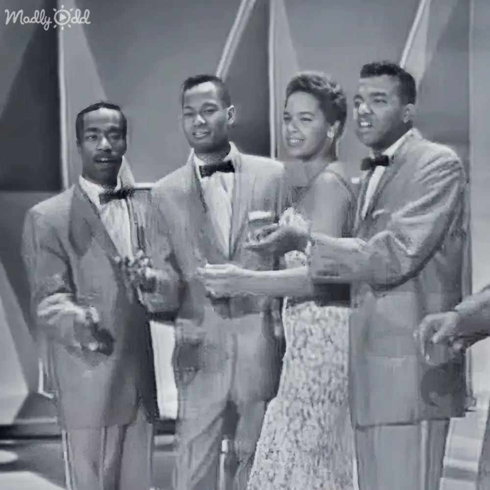 A group of young adults in formal attire performing on a modern-themed stage set against geometric backdrop.