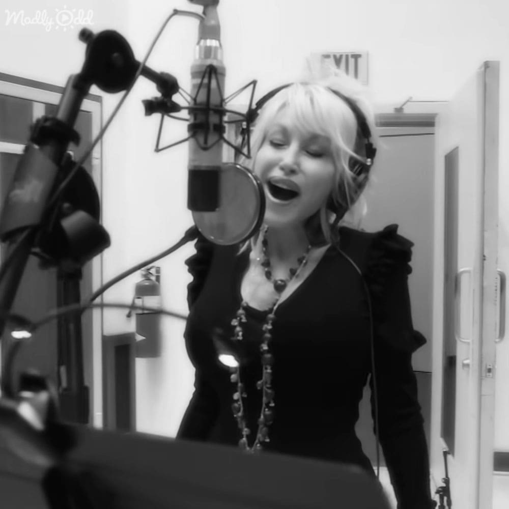 A young adult female wearing a fitted, low-cut black top with ruffled shoulders stands in an indoor recording studio setting.