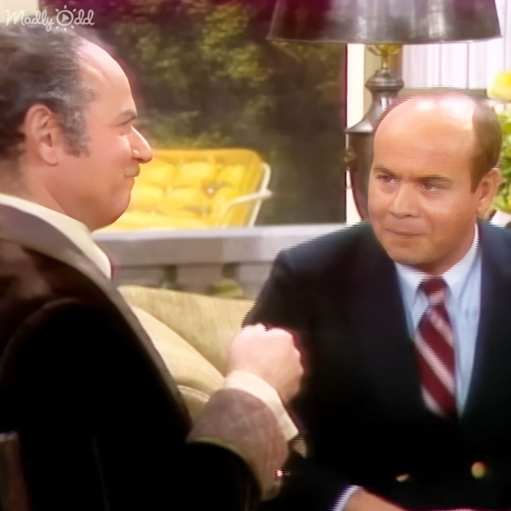 Two middle-aged men dressed formally in suits and shirts, one with a dark blazer over white shirt and another in a light blue dress shirt and red striped tie, seated indoors near yellow upholstered furniture and home decor elements.