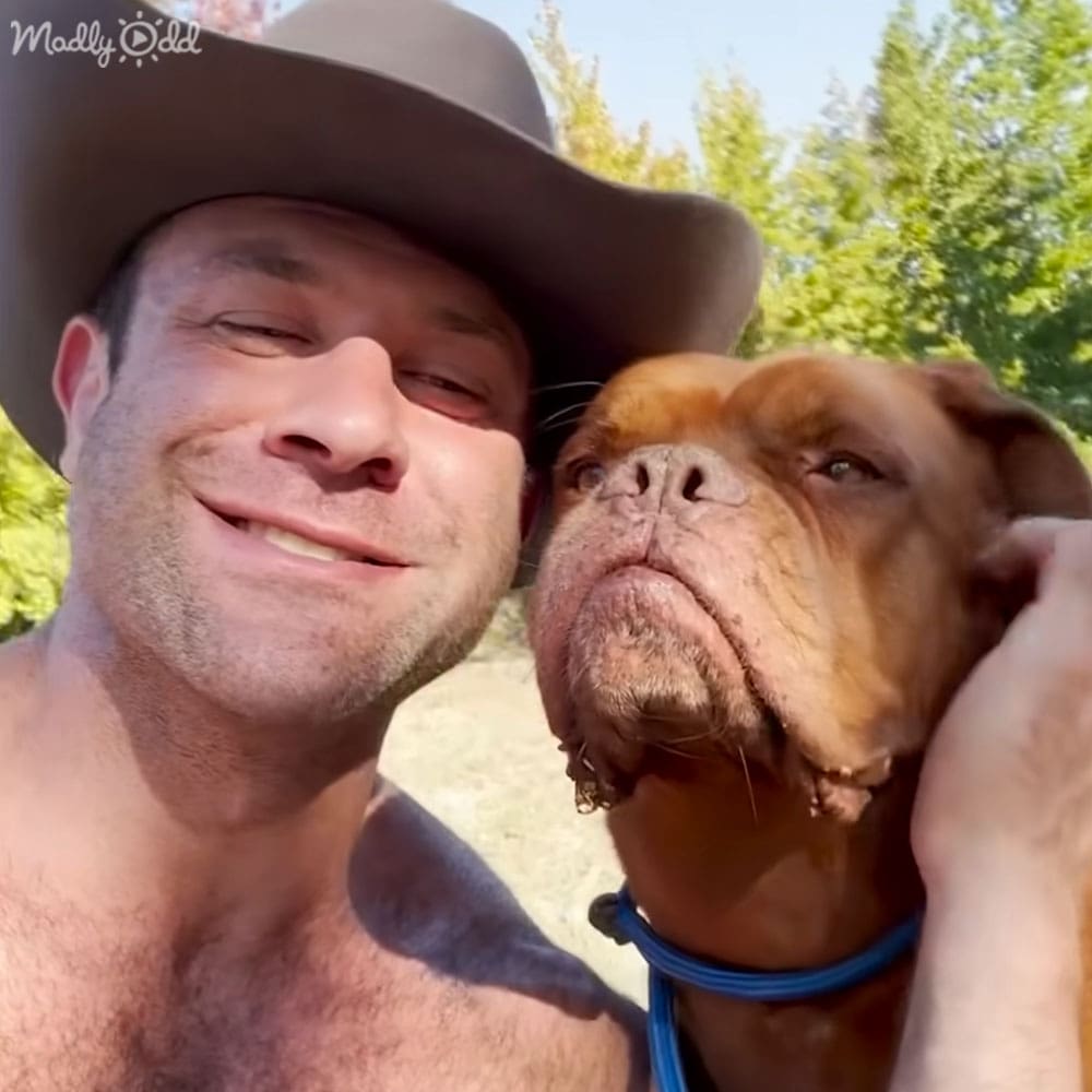 A middle-aged male wearing a wide-brimmed fabric hat outdoors with a brown pit bull dog, late spring or summer setting with clear skies and green foliage.