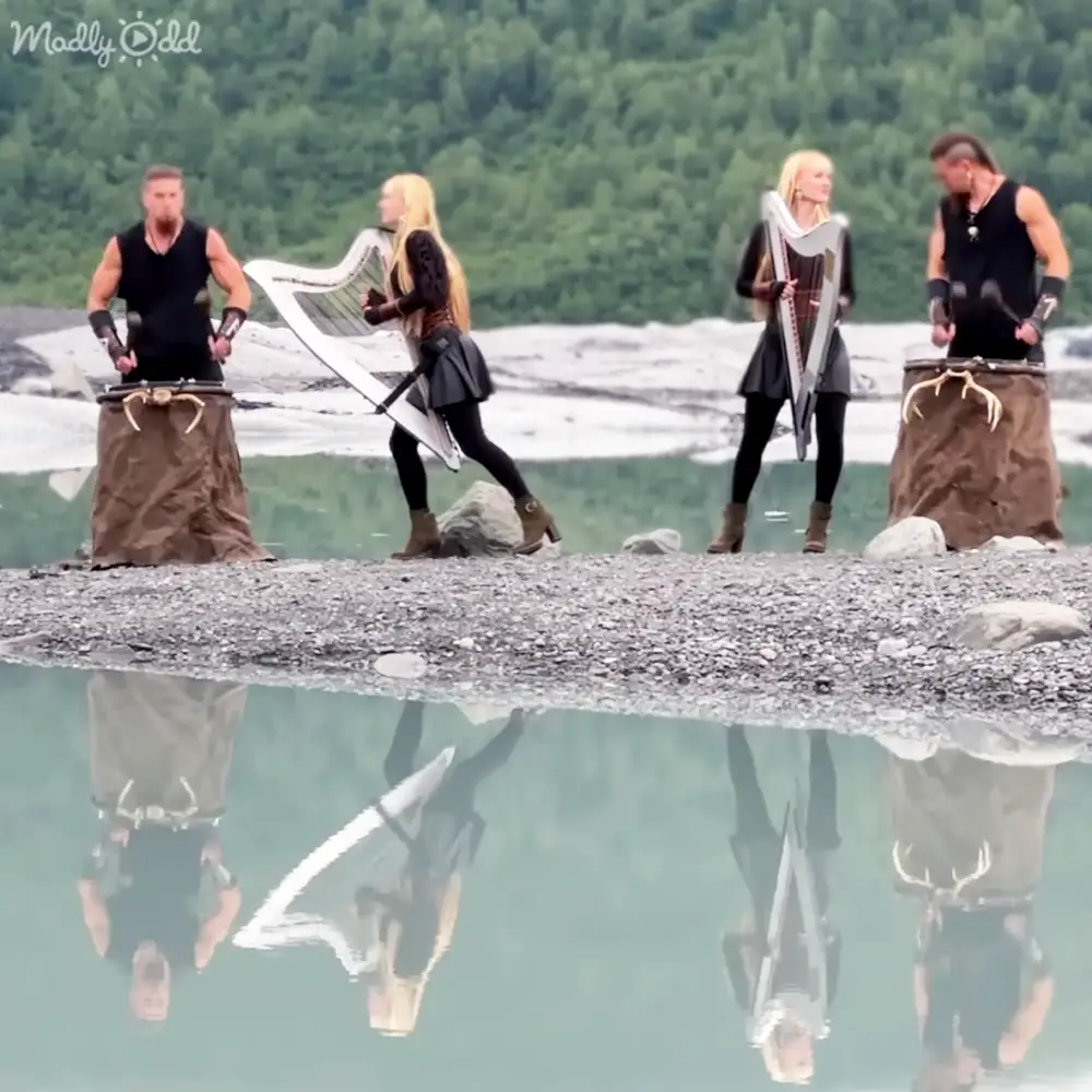 A group of adults performing music outdoors near a body of water with dense green foliage in the background, likely during daytime with overcast skies.