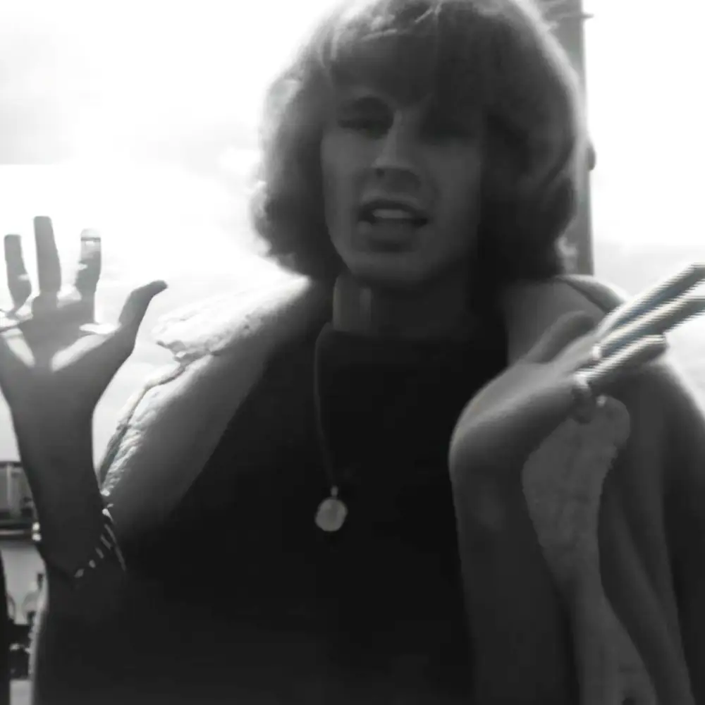 A young adult male wearing a dark-colored jacket with a high collar and fur-lined hood stands outdoors, holding what appears to be a rope or cord; black-and-white photograph suggests an urban environment.