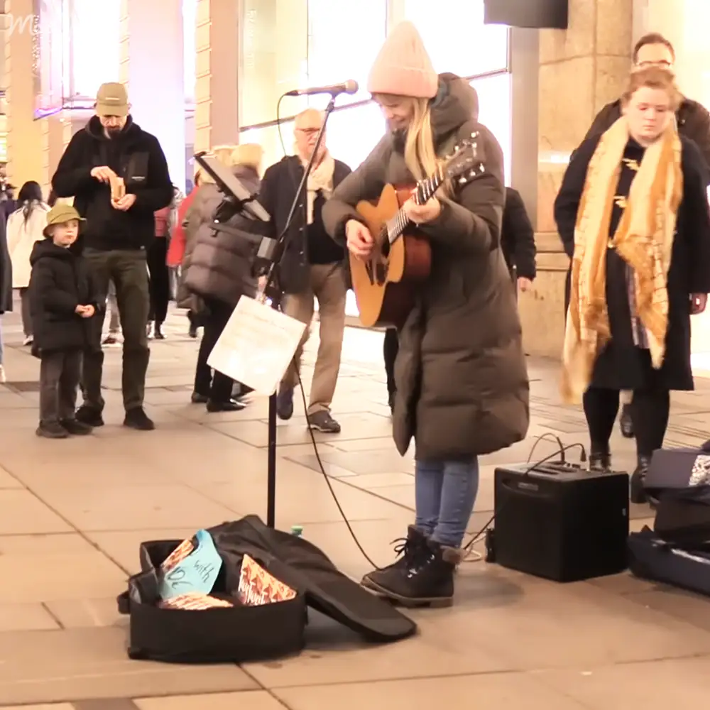 A young adult female street performer plays guitar in an urban public space with various passersby and objects around her.