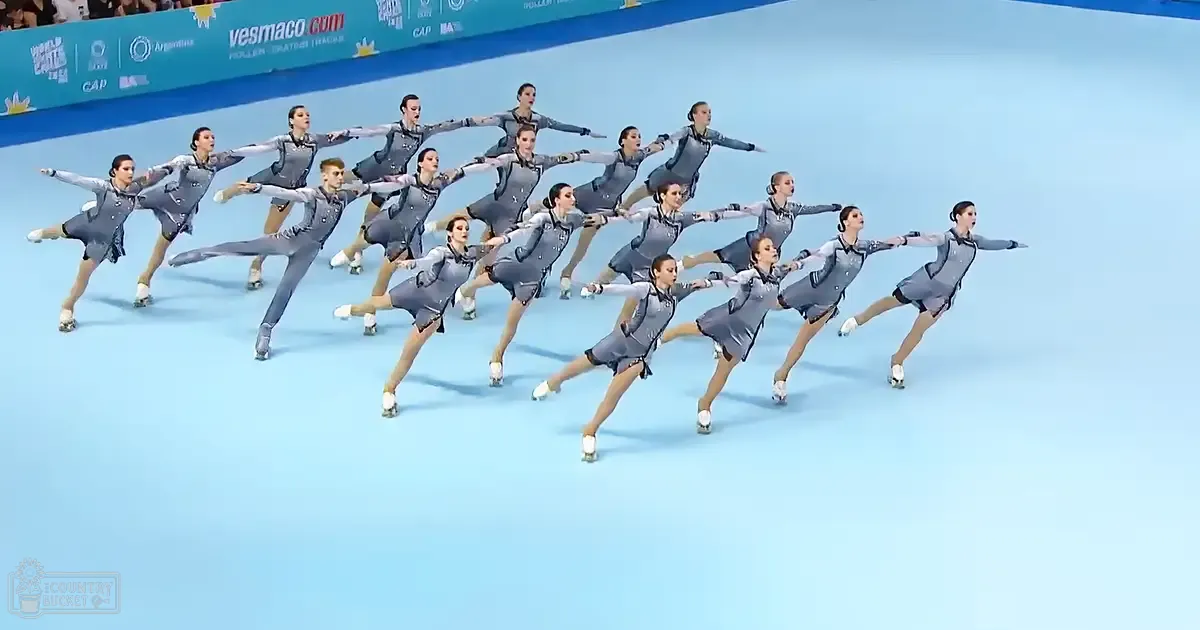 A synchronized skating team of young adult female skaters wearing gray dresses with black accents and patterns, performing at an indoor rink with light blue floors and banners related to roller skating events.