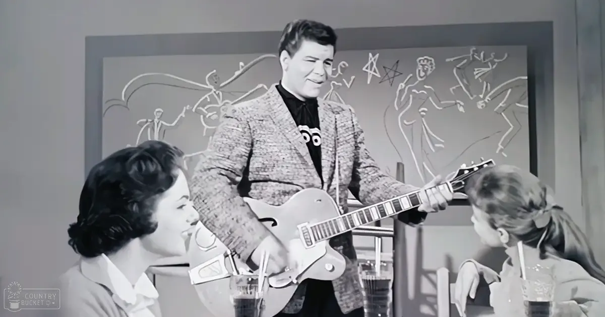 A man playing guitar indoors under dim lighting, surrounded by women; mid-20th century America ambiance.