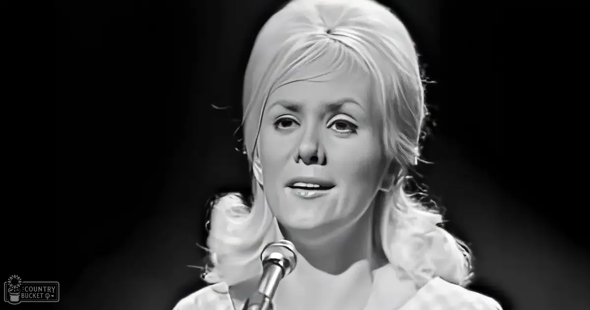 An adult female performer at a music event wearing a vintage-style white garment with ruffled details on the shoulders and sleeves; a microphone prominently featured in the foreground, possibly indicating a concert or recording session.