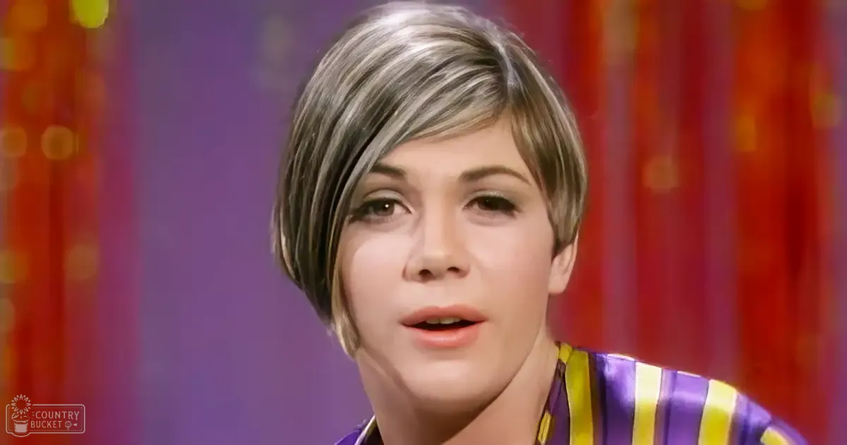 A young adult female wearing a striped top stands against a colorful, vertically striped background with red and purple hues. The setting appears to be indoors, likely for an interview or talk show segment.