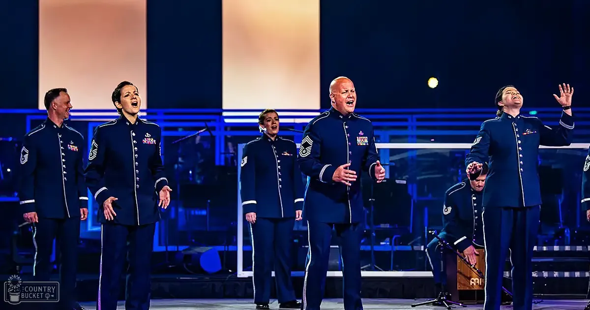 A group of adult male and female performers in uniform military-style attire, likely at an indoor concert or event setting with stage lighting and musical instruments visible.