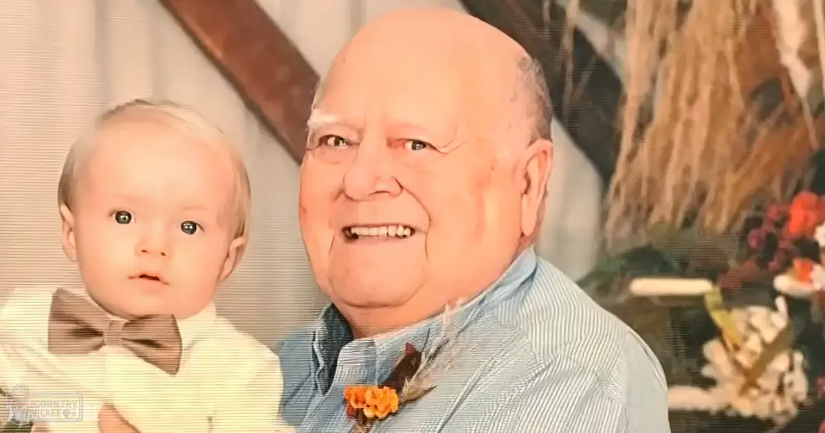 An infant in a white shirt with a brown bow tie held by an older adult wearing a blue and white striped button-up shirt with a patterned flower, set against the backdrop of a cozy home interior with floral arrangements and personal decor.