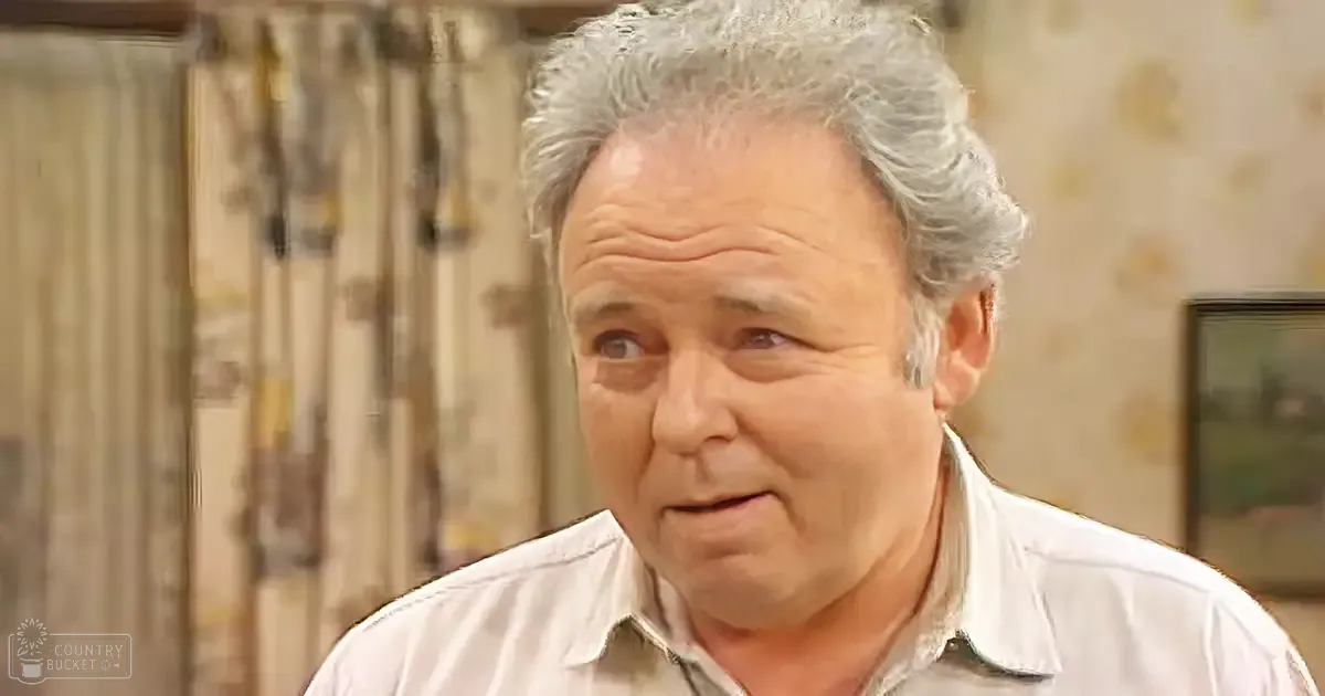 Middle-aged to older adult male wearing a light-colored collared shirt indoors with natural daylight streaming through curtains.