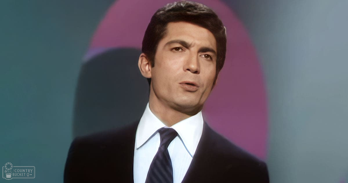A man in a classic tailored suit with dark jacket, white shirt, and dark tie stands indoors. The setting suggests an indoor performance space or studio with controlled lighting.