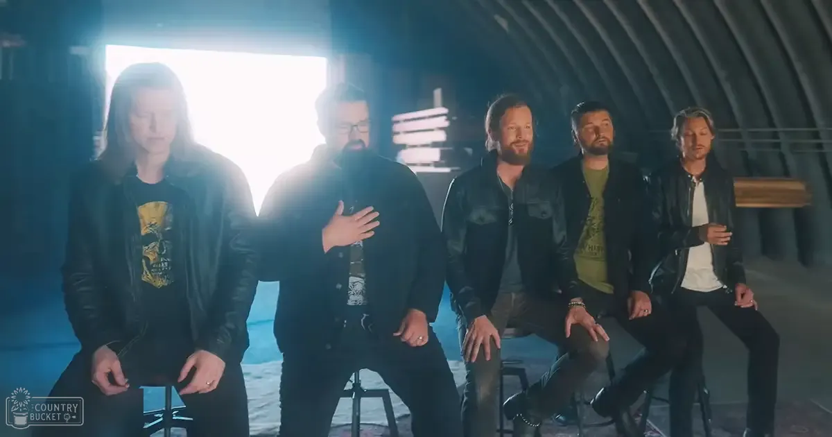 Five adults seated indoors under dramatic lighting, wearing casual and graphic t-shirts, leather jackets, and dark pants in a space with industrial elements like metal beams and black stools.
