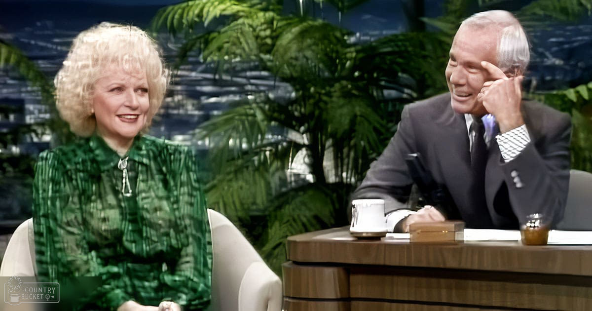 A talk show host in a green patterned blouse interacts with an older male guest dressed in a dark suit. The setting is an indoor studio with plants, desk items, and a scenic backdrop.