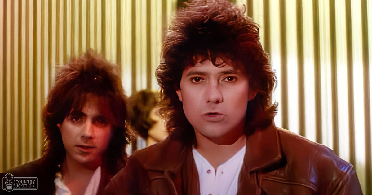 A young adult male in a brown leather jacket and white shirt stands indoors near another similarly dressed individual, suggesting a casual or professional setting.