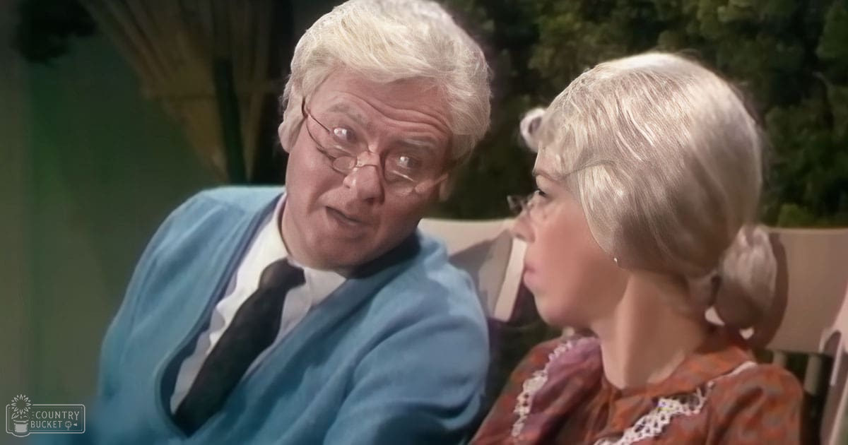 An older adult male in a light blue cardigan over a white shirt with a dark tie and glasses, seated outdoors with an older adult female wearing an orange patterned top with lace detailing in a lush garden setting under natural daylight.