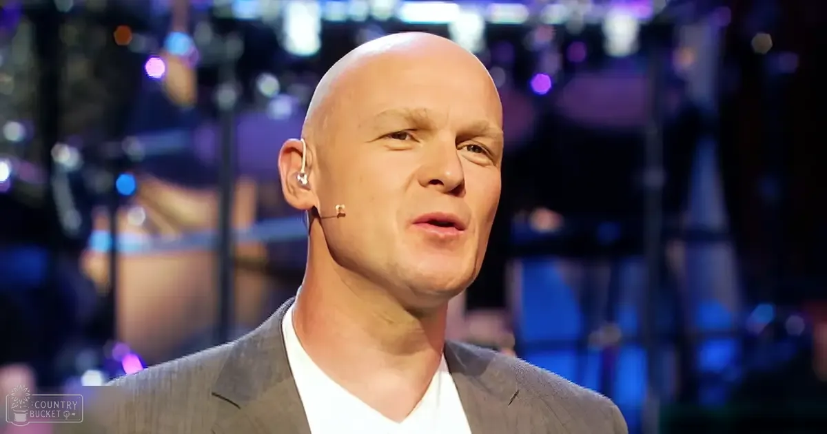 A middle-aged adult male in a tailored gray suit jacket and white collared shirt stands on what appears to be a stage or studio with blurred background lights suggesting an event, performance, or presentation.