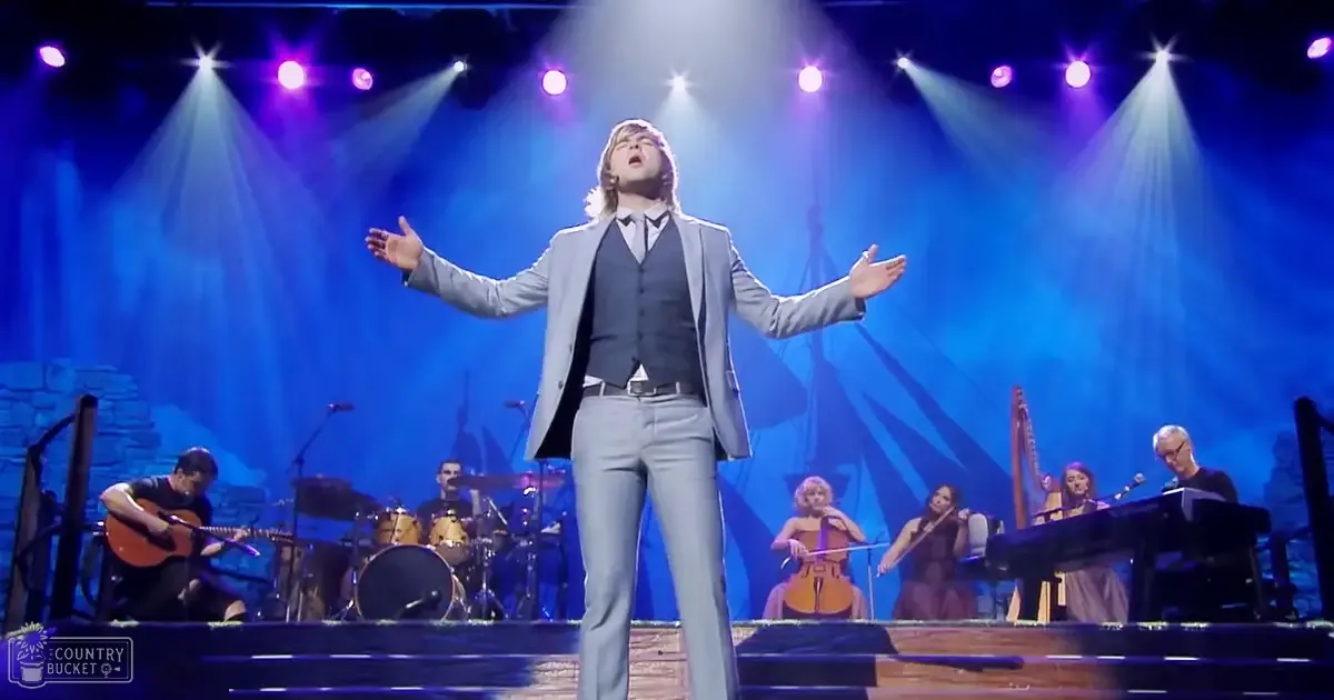 A young adult male singer performing on stage at an indoor concert venue with musicians and a stone backdrop under blue and purple stage lighting.