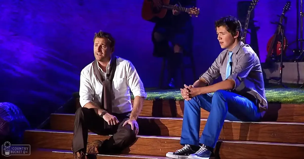 Two young adult males performing on a stage with blue and purple lighting, seated on wooden benches; one wearing a white button-up shirt and brown scarf, another in a light-colored shirt and black sneakers.