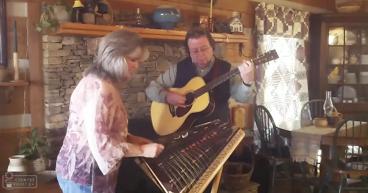 A musical performance by an older adult female wearing a tie-dye patterned shirt indoors in a cozy home setting with wooden chairs, a stone fireplace, and rustic decor.