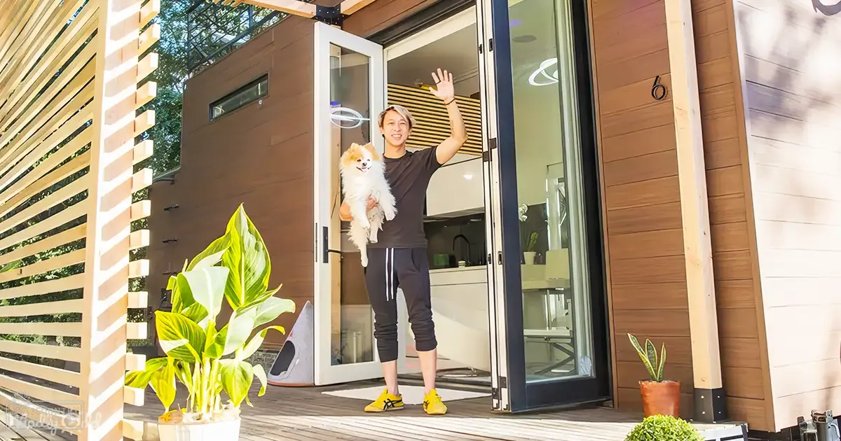 A young adult male stands outdoors at a modern house with sliding glass doors, dressed in dark gray t-shirt and black athletic pants with white stripes, yellow sneakers. A small Pomeranian dog with white coat and light brown markings is nearby.