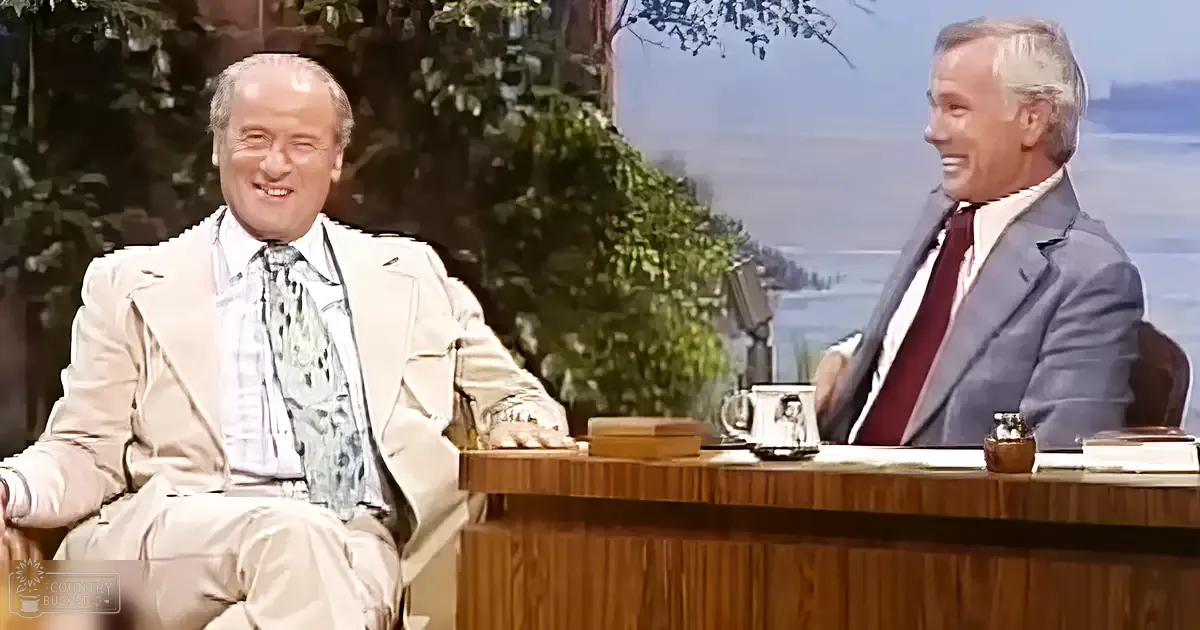 Talk show host and guest seated on a wooden desk with greenery outside, suggesting spring or summer in a well-lit studio setting.