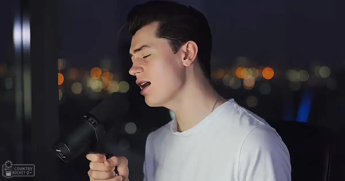 Young adult male singer wearing a white t-shirt with short sleeves, holding a microphone against an urban backdrop at night.