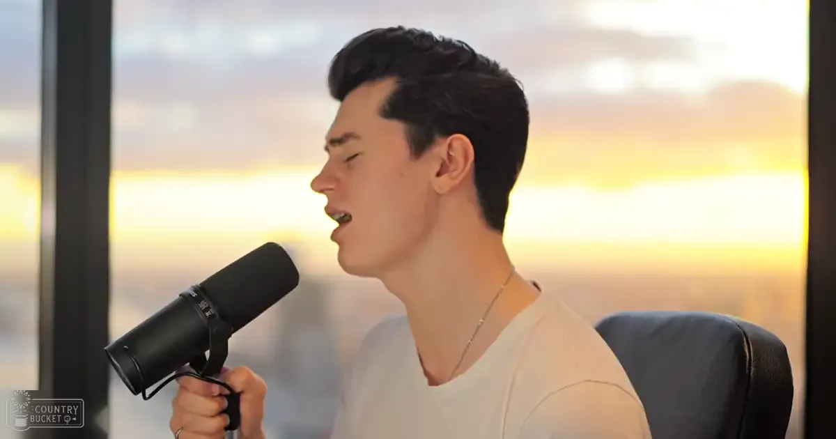 A young adult male wearing a white, relaxed casual t-shirt sings into a microphone with a black pop filter attached. He sits on a chair in an indoor space with large windows showing an outdoor view of a sunset or sunrise.