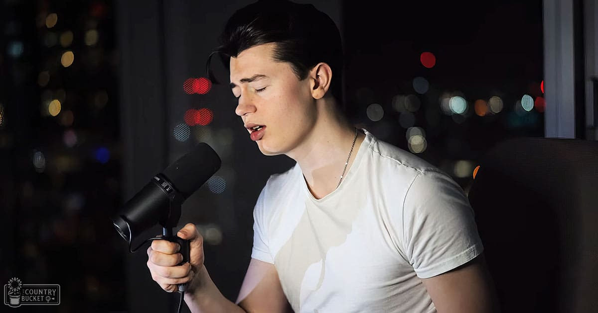 A young adult male singer wearing a white t-shirt with relaxed fit and V-neck holding a black microphone, set against city lights at night.
