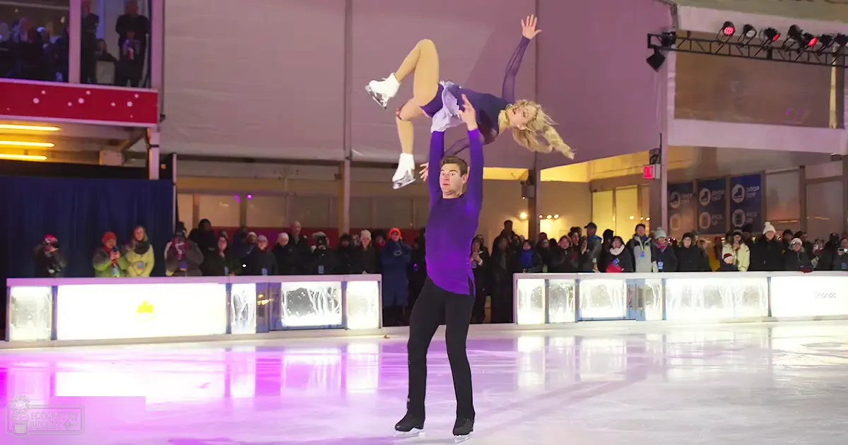 An adult male skater in a long-sleeved purple top and black pants performs at an indoor ice rink with spectators watching; evening or night setting, winter attire visible.