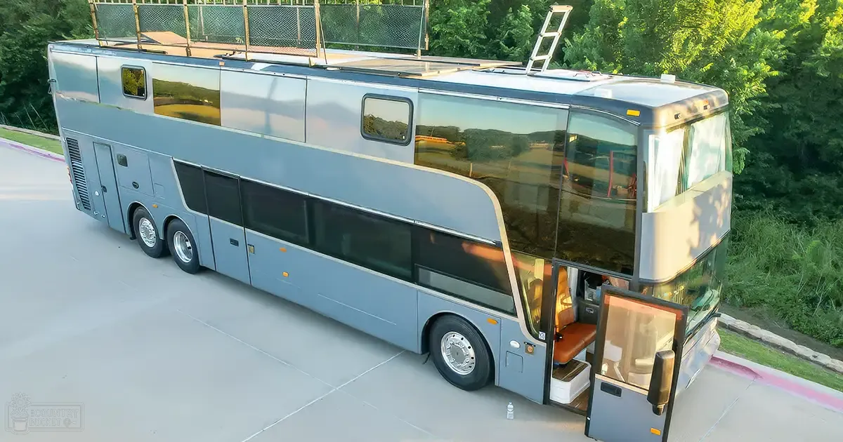 A modern double-decker bus repurposed as a mobile home, surrounded by trees and green foliage on a paved area during daytime with clear weather conditions.