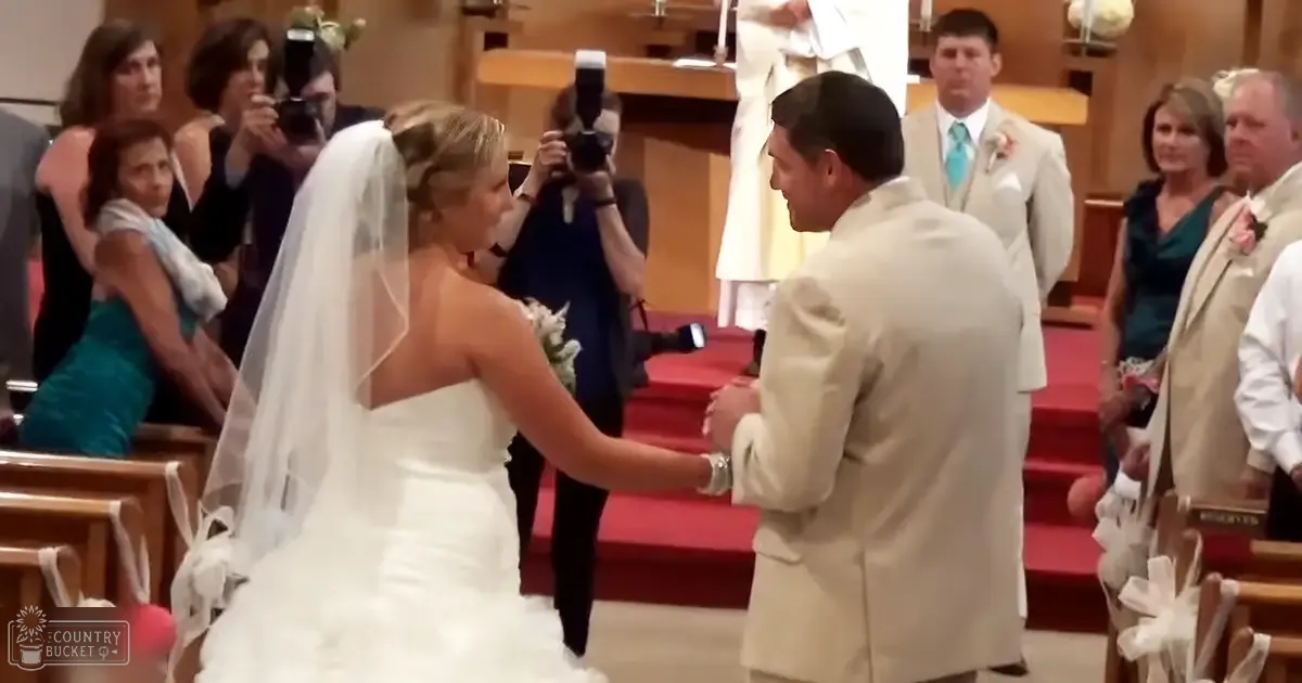 A bride and groom at a wedding ceremony inside a church, with red carpeted steps leading to an altar adorned with white floral arrangements under natural daylight.